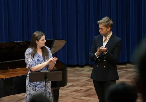 Two people, a man and a woman, standing in front of a blue curtain on a stage with a piano. The woman, wearing a floral dress, is speaking and gesturing with her hands. The man, dressed in a dark blazer and white shirt, is holding a piece of paper an