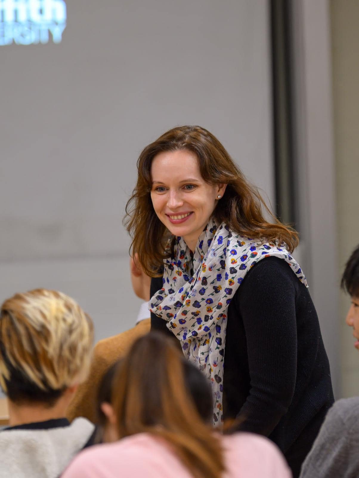 A woman with shoulder-length brown hair, dressed in a black top and a white scarf with colorful floral patterns, smiling and engaging with a group of people in an indoor setting.