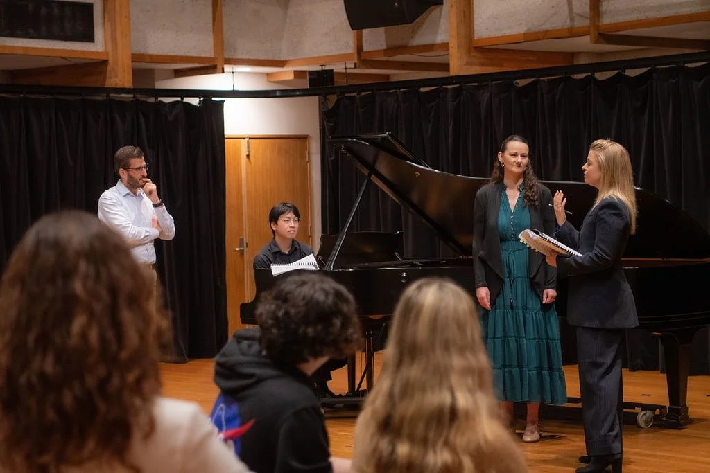 Two women are standing and talking near a grand piano, while a man stands to the side listening at a music rehearsal or performance, in a room with black curtains and wood accents. Several audience members are visible in the foreground.