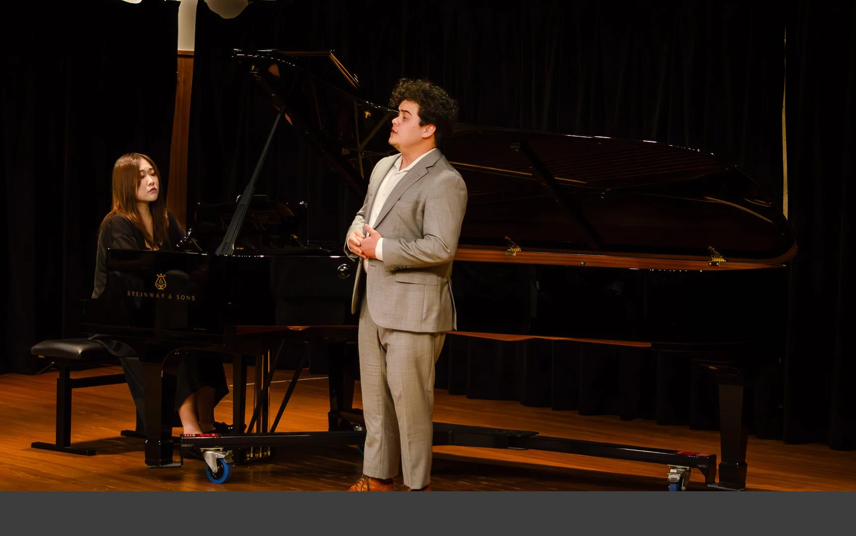 A man in a beige suit standing on stage in front of a black grand piano with a woman sitting at the piano, performing or practicing, against a black curtain background.