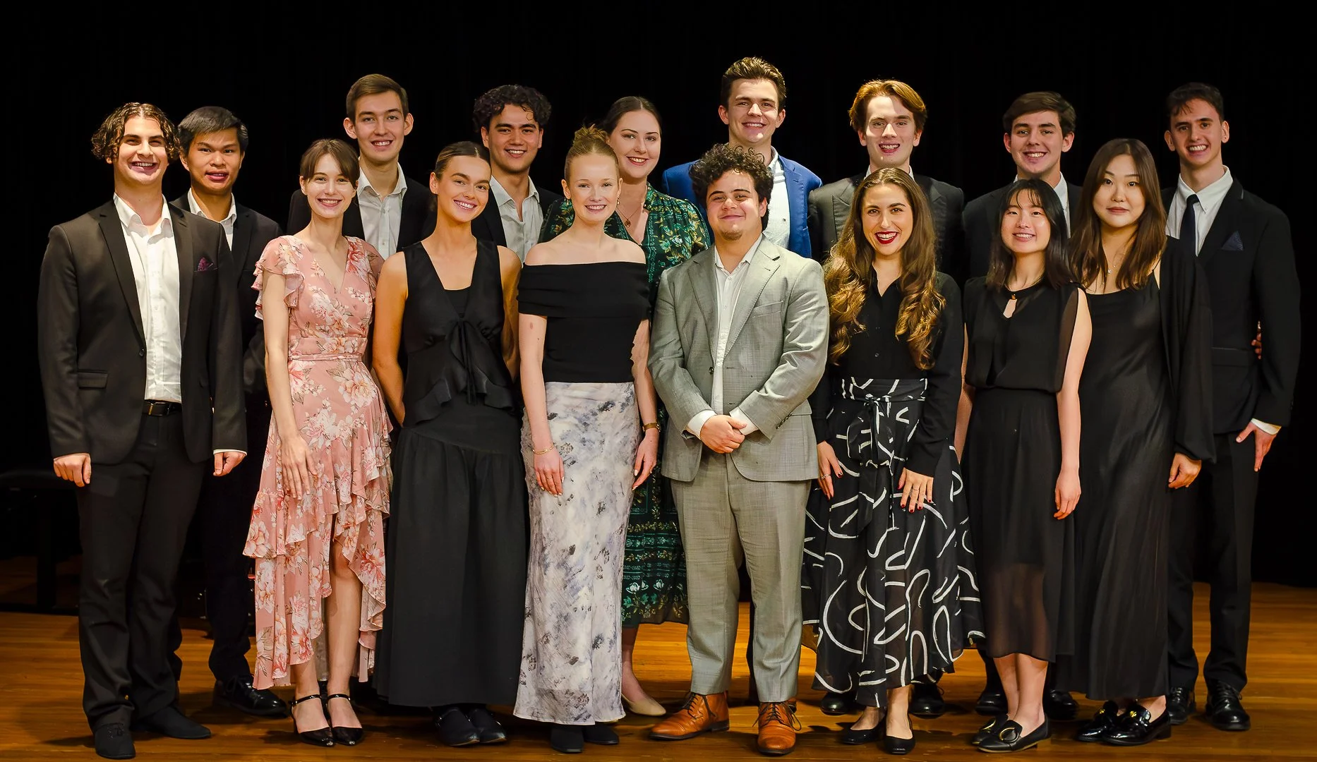 Group of young adults dressed in formal attire, standing on a stage with a black background, smiling for a photo.