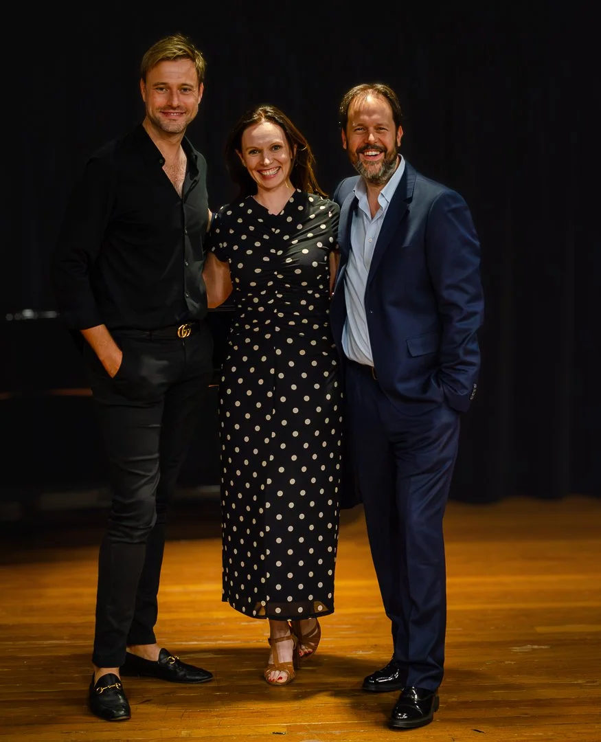 Three people standing together on a wooden floor against a black backdrop, smiling at the camera. The man on the left is wearing a black shirt and pants with loafers, the woman in the middle is wearing a black dress with white polka dots and high hee