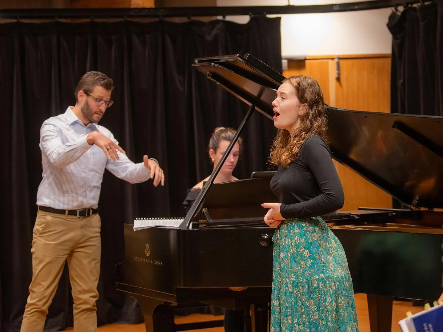 A woman singing at a piano during a rehearsal, with a conductor guiding her and another musician playing a piano in the background.