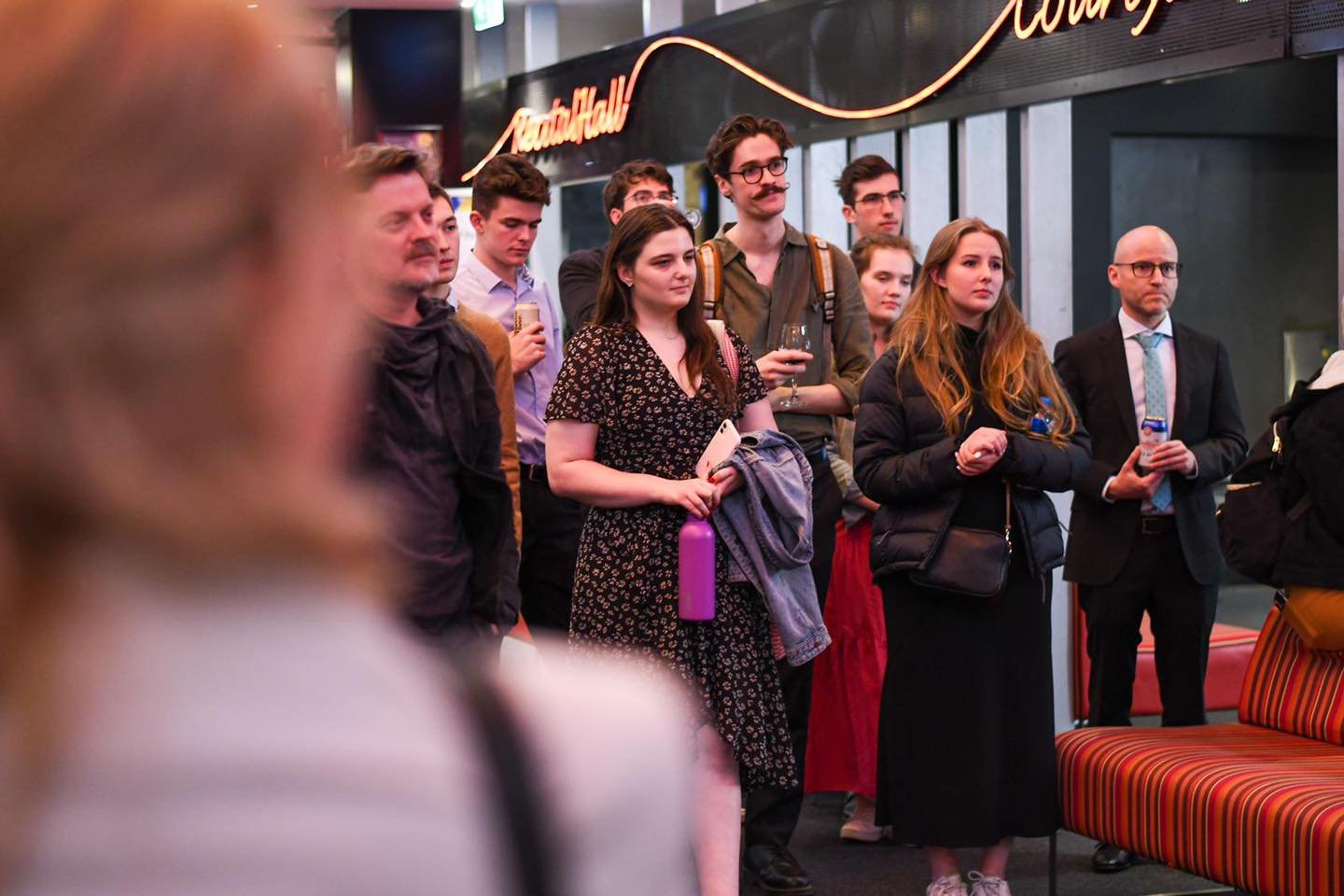 A group of people standing indoors, listening attentively, some holding drinks and personal items, with a neon sign and dark decor in the background.