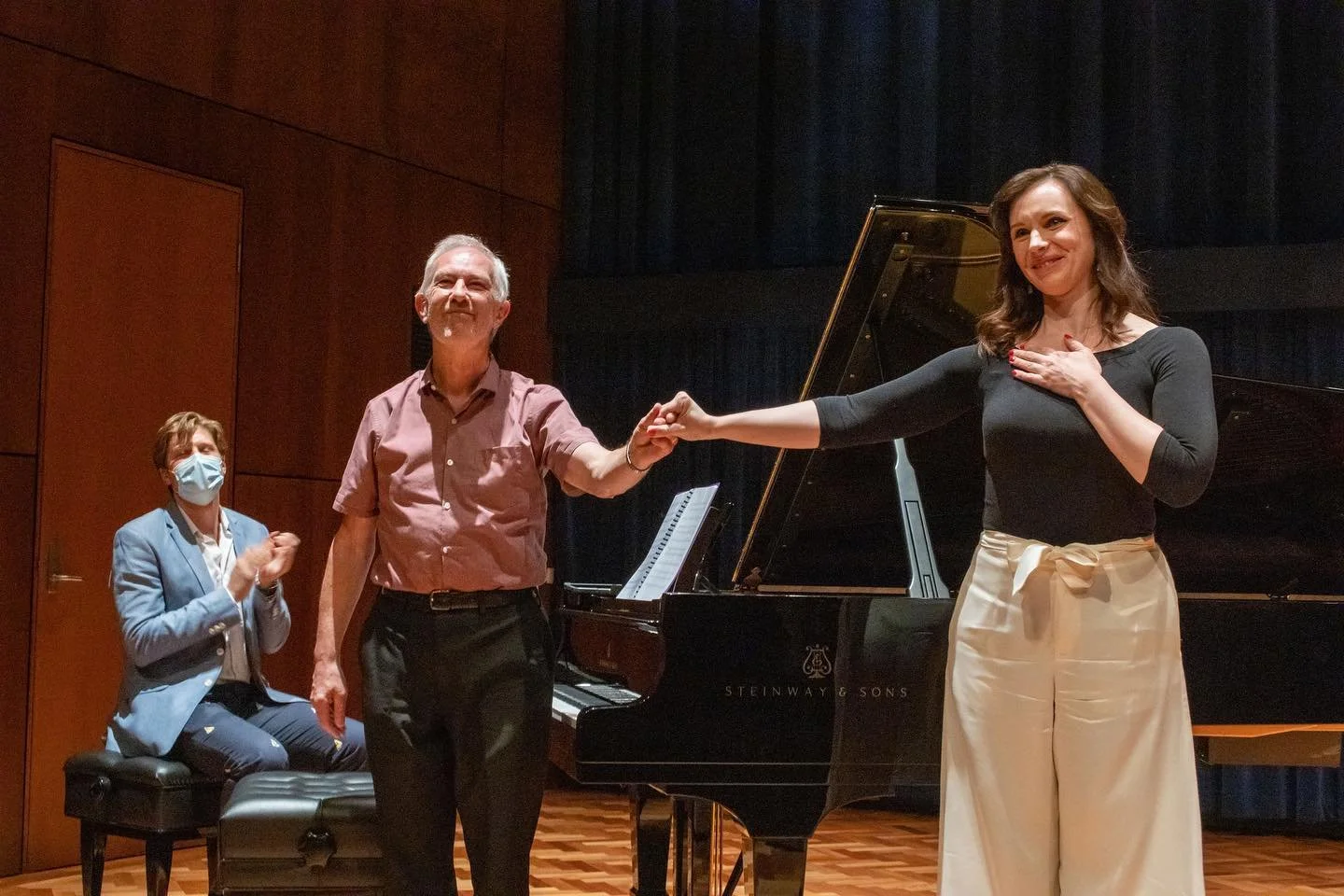 A woman receiving an award or recognition from an older man at a piano recital or concert hall, with another woman clapping in the background.