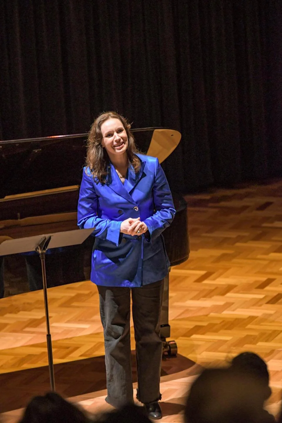 A woman in a bright blue blazer and dark pants standing on a wooden stage, smiling near a grand piano and a music stand, with a black curtain in the background.