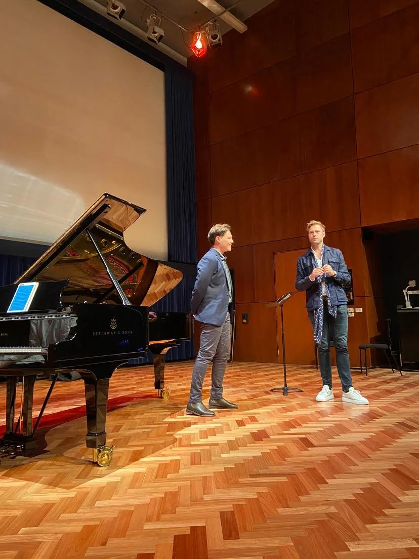 Two men stand on a stage in a music rehearsal or performance space, one is wearing a blazer and the other a denim jacket, with a grand piano nearby.