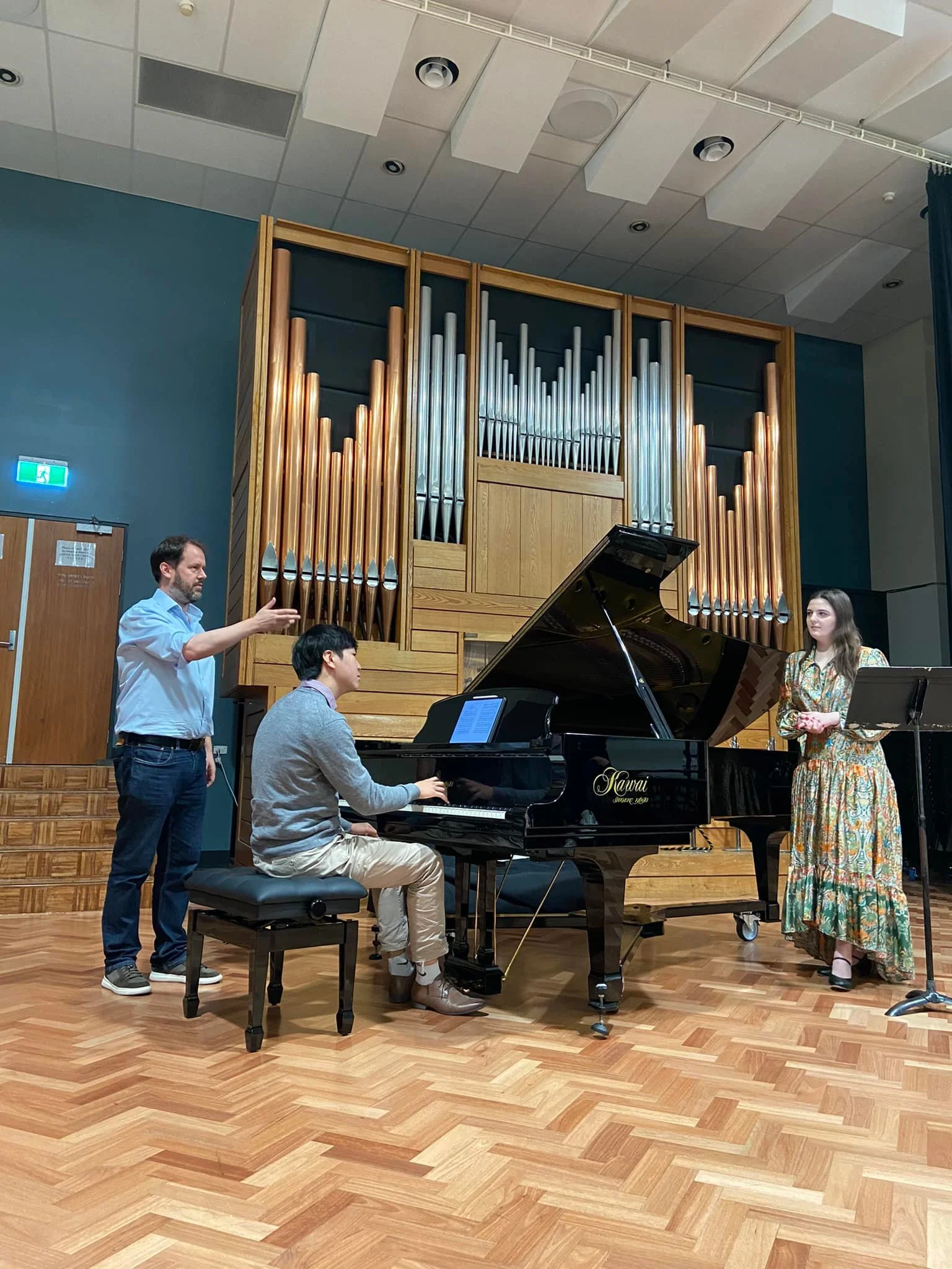 A music rehearsal in a concert hall with three people: a man standing on the left, a man playing a piano, and a woman standing on the right. The hall features large pipe organ pipes in the background.