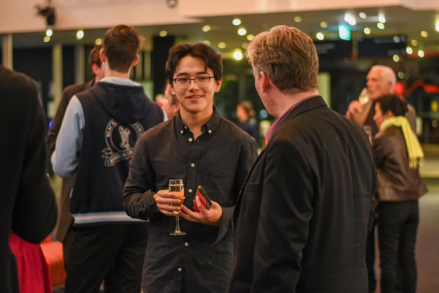Two men engaged in conversation at a social event, one holding a champagne flute, in a lively, warmly lit indoor setting with other people mingling in the background.