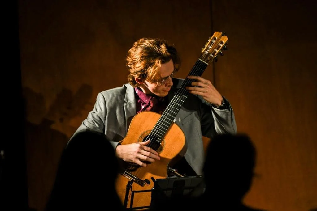 A man with wavy brown hair in a suit playing classical guitar on stage, with audience silhouettes in the foreground.