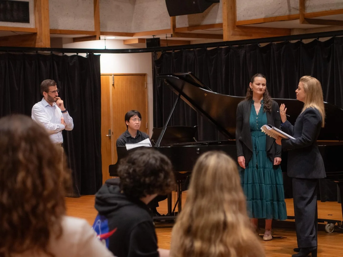 A panel discussion or interview with five people on stage, including a woman in a teal dress and black jacket, a woman in a gray suit holding a notepad, a man in a white shirt with glasses, and a musician sitting at a grand piano, with an audience in