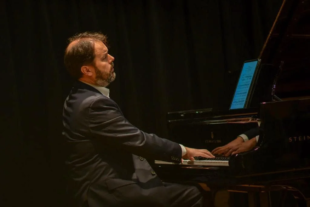 A man with a beard in a suit playing a grand piano on a stage with black curtains.