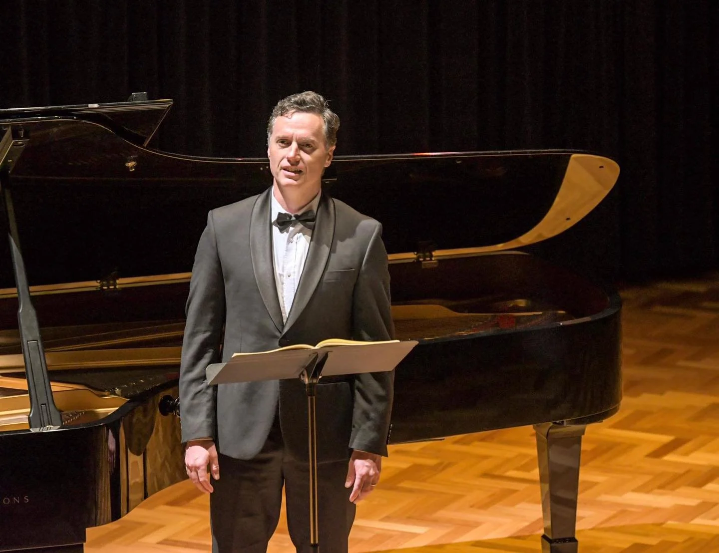 Man in formal tuxedo standing in front of a grand piano on a stage with a music stand.