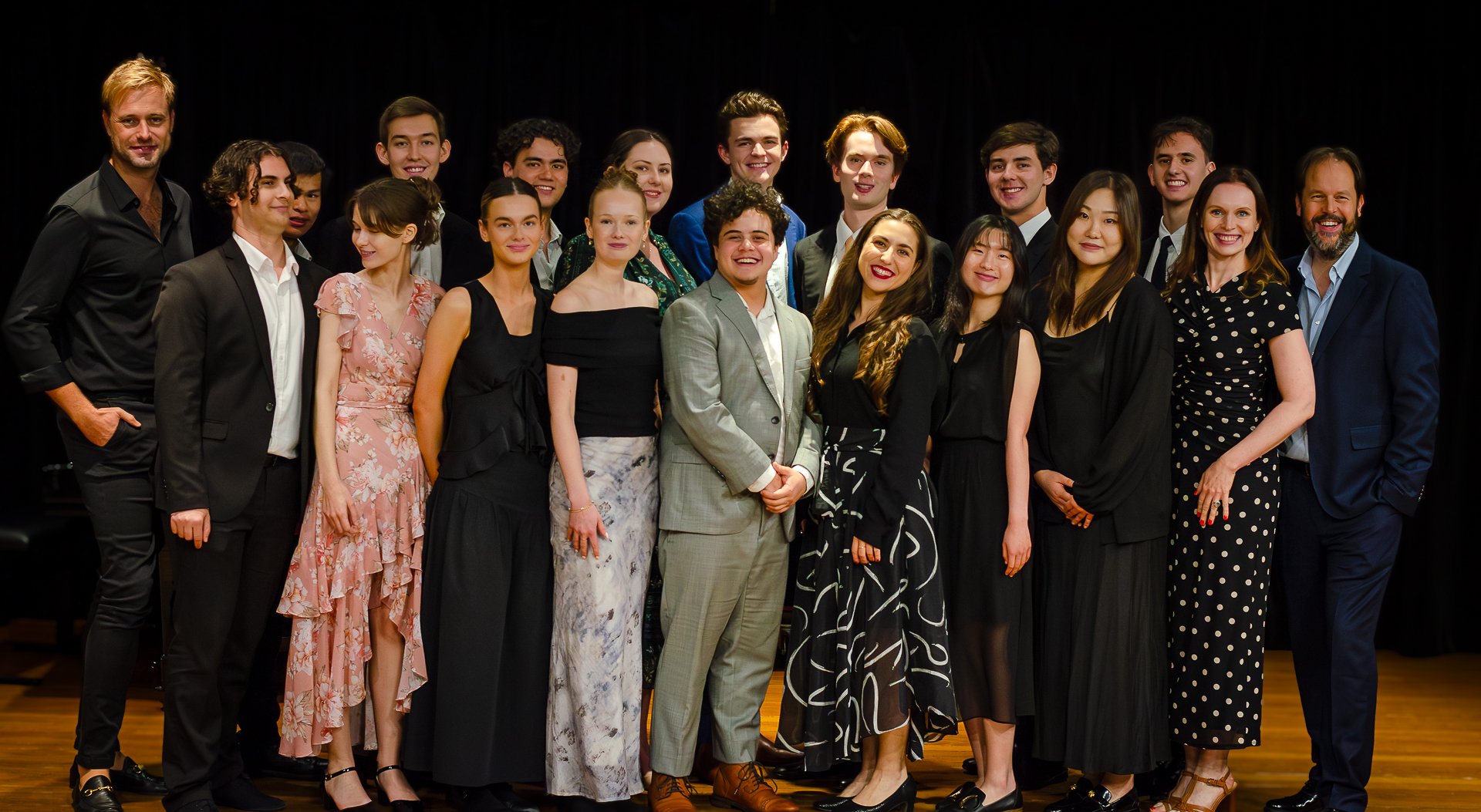Group of young performers and two adults posing on stage with a black curtain backdrop.
