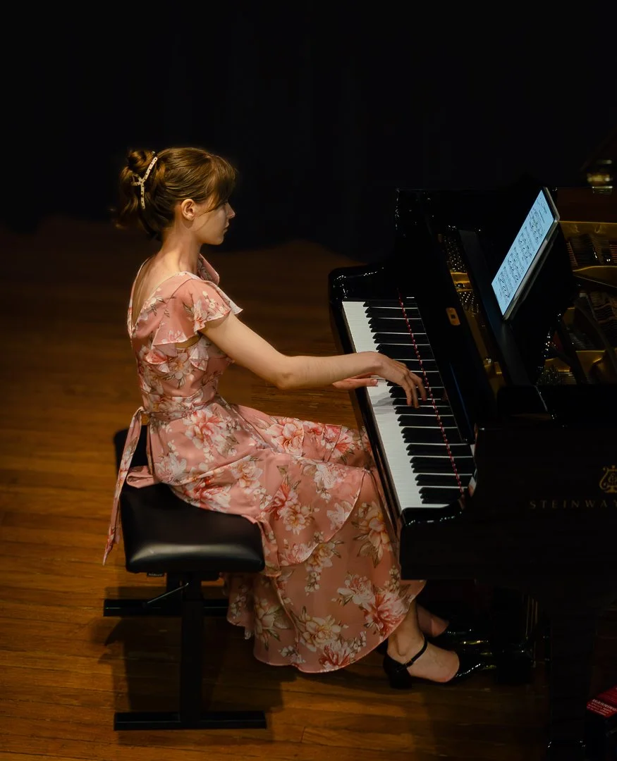A woman in a pink floral dress playing a grand piano on a wooden stage.
