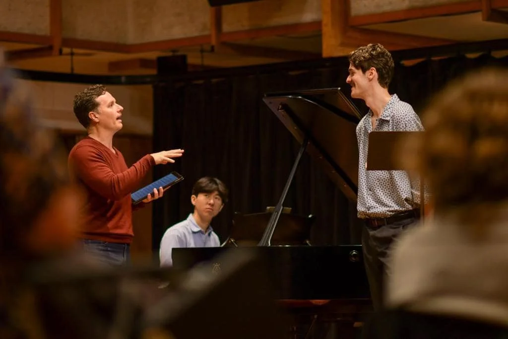 A woman appears to be conducting a musical rehearsal or class, engaging with a man at a grand piano and a young boy seated at the piano, in a room with wooden beams and a black curtain.