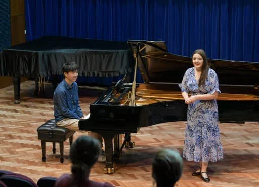 A young man playing the piano and a young woman singing or speaking on a stage in front of an audience, with a grand piano, blue curtain backdrop, and wooden floor.