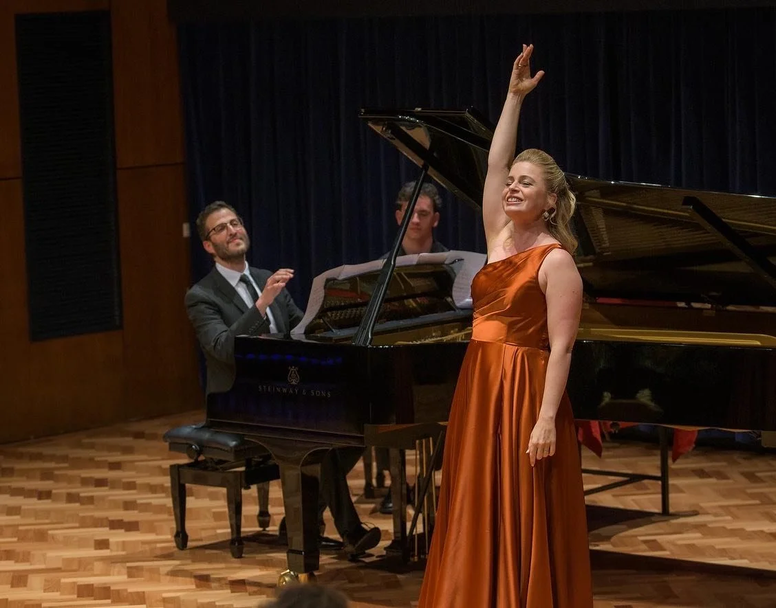 A woman in an orange satin gown singing on stage with a pianist and another musician in the background.