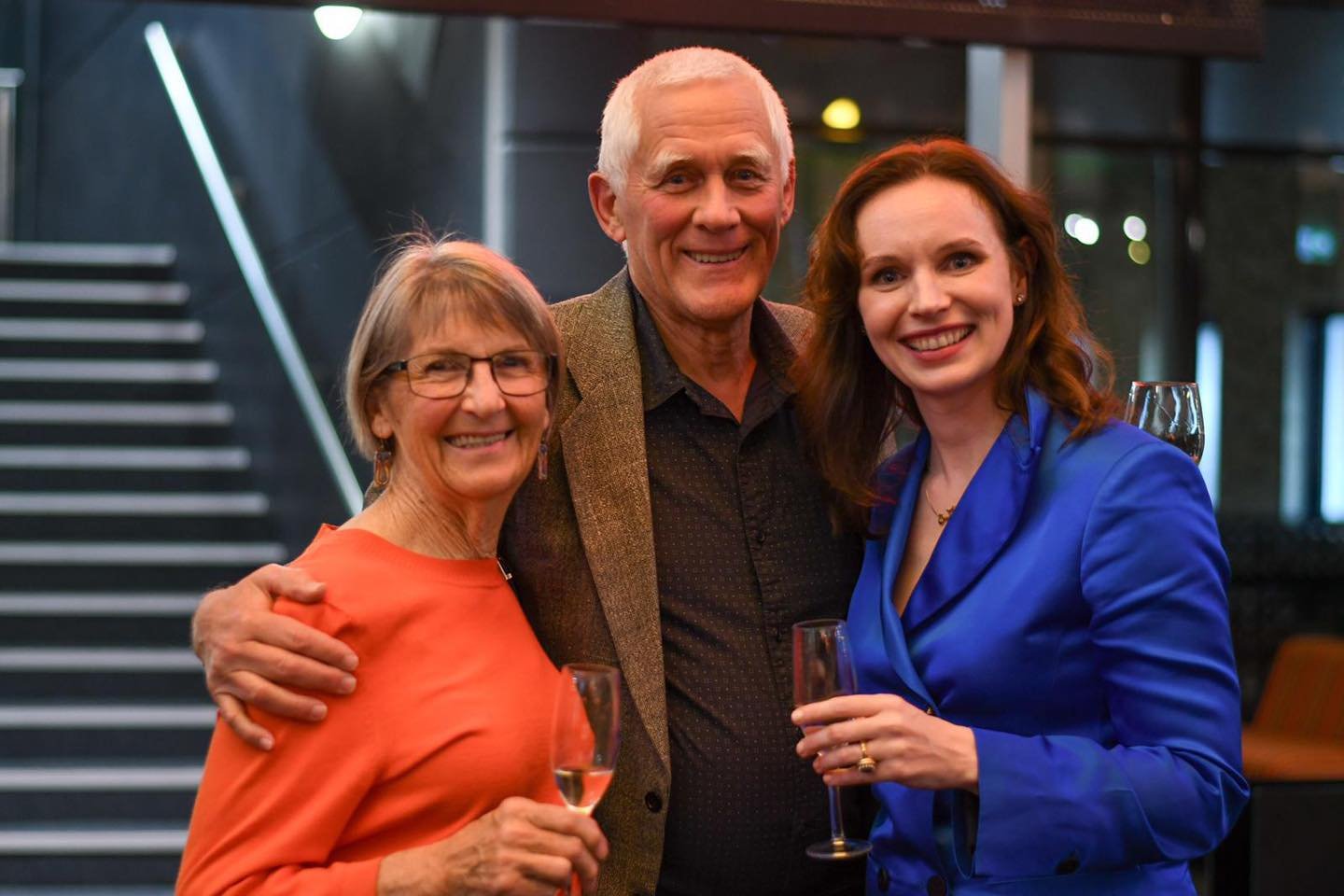 Three people at a social gathering, two women and one man, smiling and holding glasses of champagne, in an indoor setting with modern decor and staircase in the background.