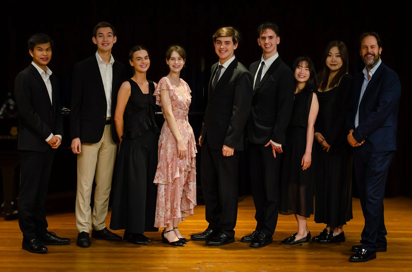 Group of ten people dressed formally standing on a wooden floor in front of a dark background.