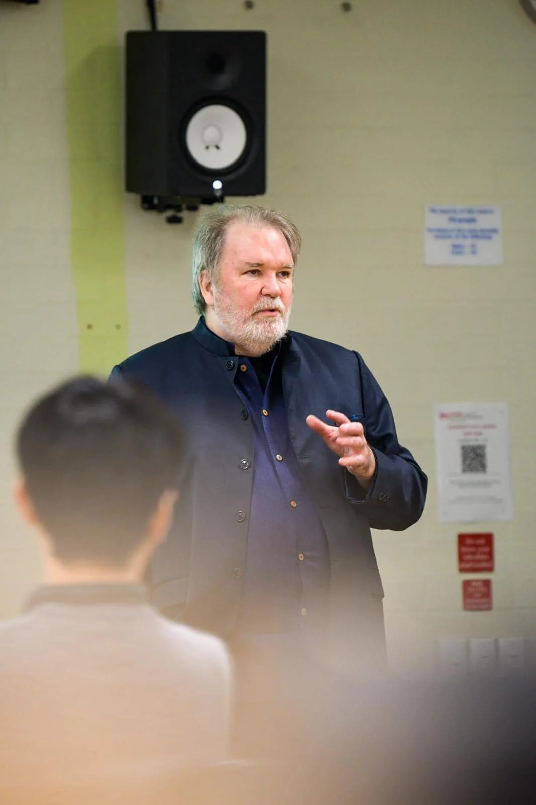 A man with gray hair and a beard speaking to a group, with a speaker mounted on the wall behind him and signs on the wall.