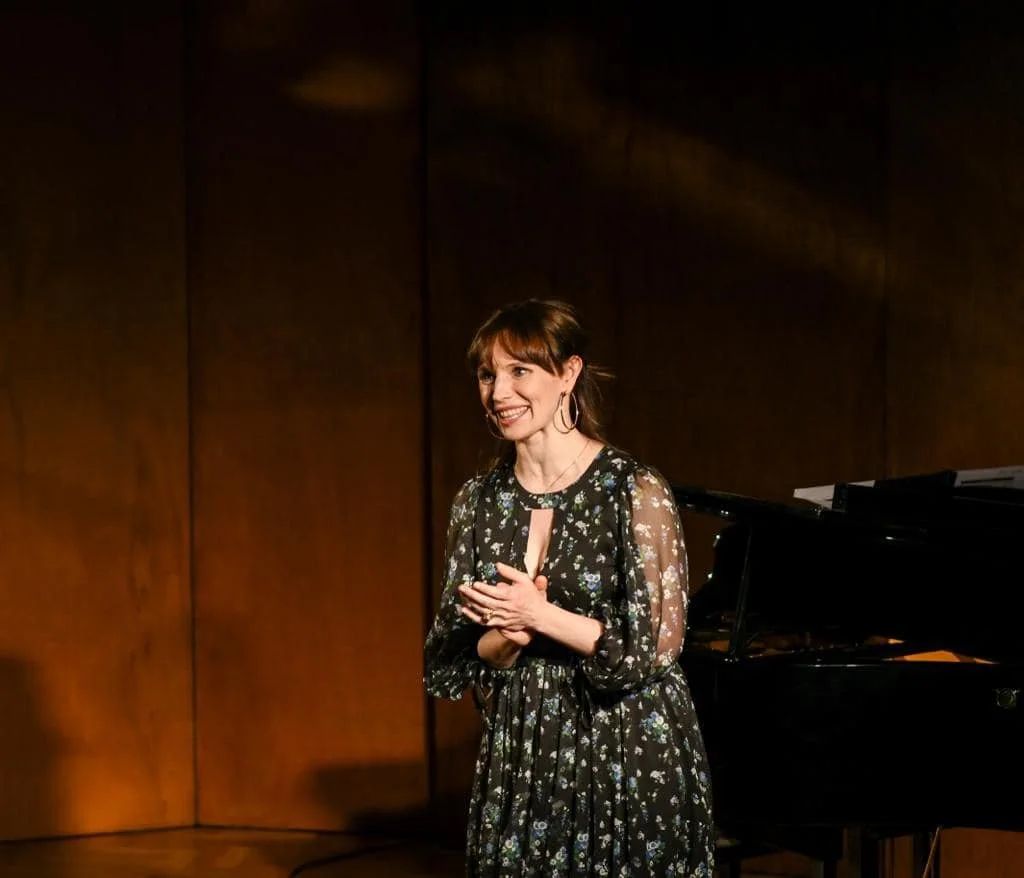 A woman in a black floral dress standing in front of a grand piano, smiling with her hands clasped, in a wooden-finish room.