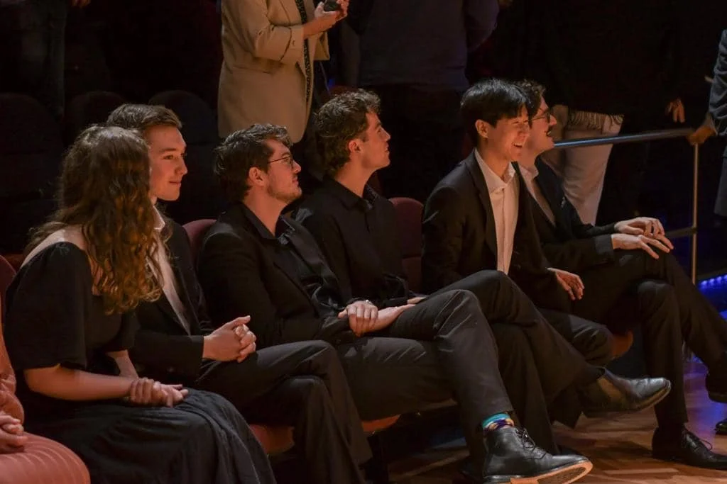 Group of young adults seated in a dark auditorium, watching a performance with smiles and expressions of enjoyment.
