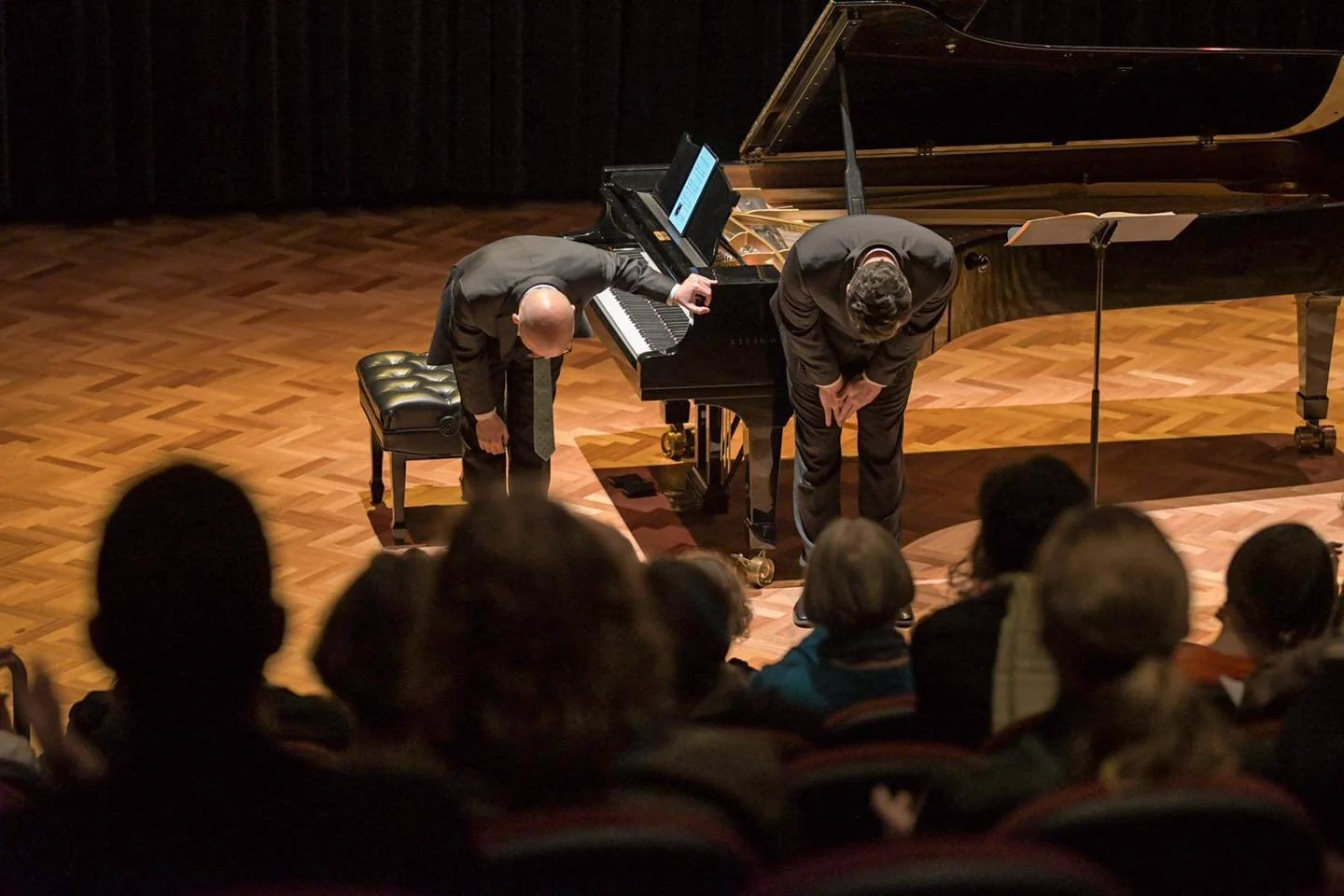 Two musicians bow to the audience on a wooden stage with a grand piano in a concert hall.