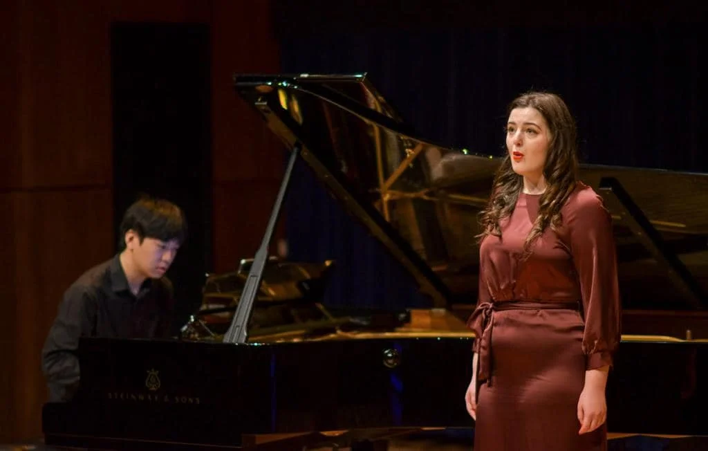 A woman singing at a concert with a man playing the grand piano.