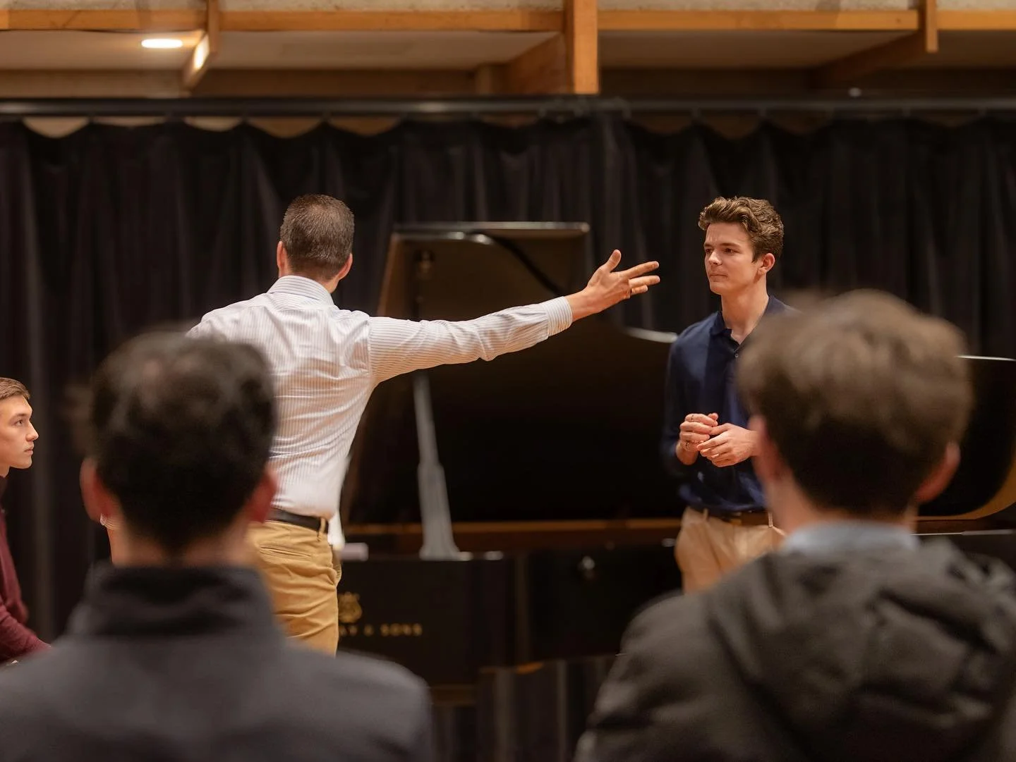 A music class or rehearsal with a young man in a dark shirt standing near a grand piano, a teacher or conductor gesturing with his arm extended, and other students seated nearby, in a room with black curtains and wooden beams.
