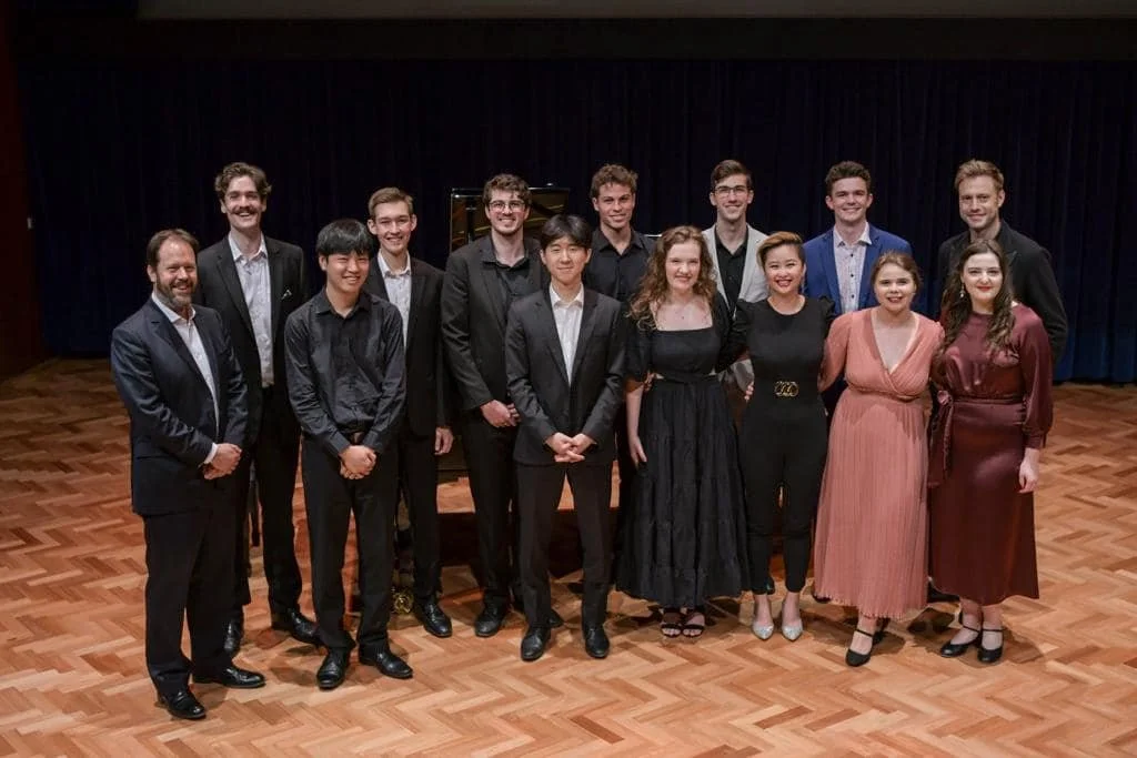 Group of 13 young adults dressed in formal attire, standing on a wooden stage with a dark curtain background, posing for a photo.
