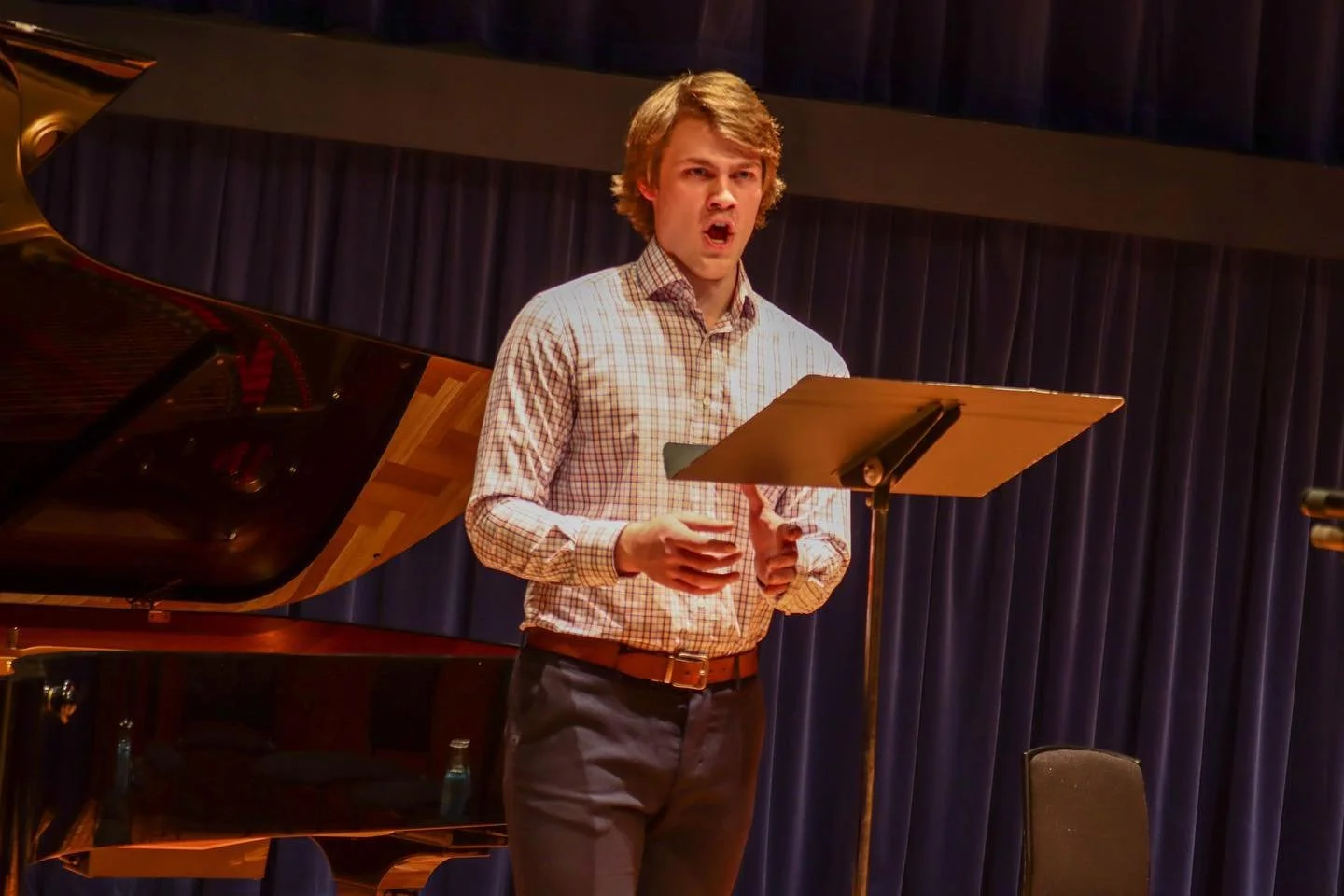 A young man in a checkered shirt singing or performing on a stage in front of a blue curtain, with a grand piano beside him.