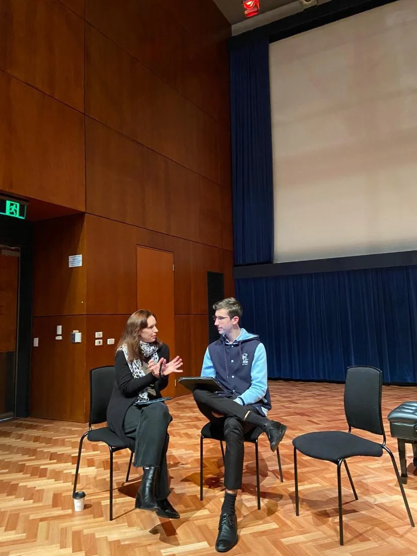Two people sitting on chairs in a room with wooden walls and a large blank screen, engaged in conversation.