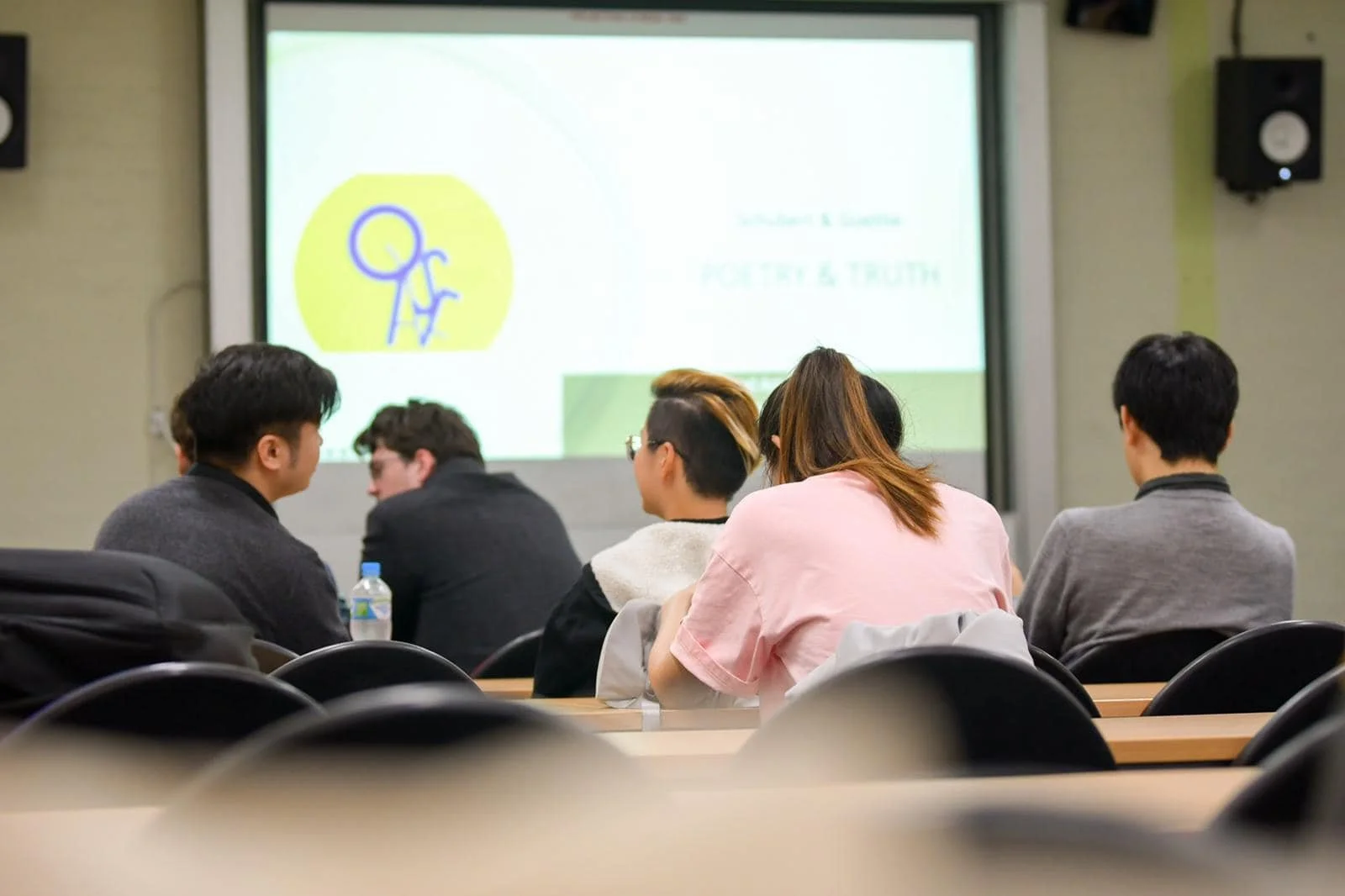 A group of people sitting in a classroom or conference room, facing a large projection screen at the front. The room has black chairs and some tables, with a water bottle on one table. The screen displays a logo of a person with a backpack and some t