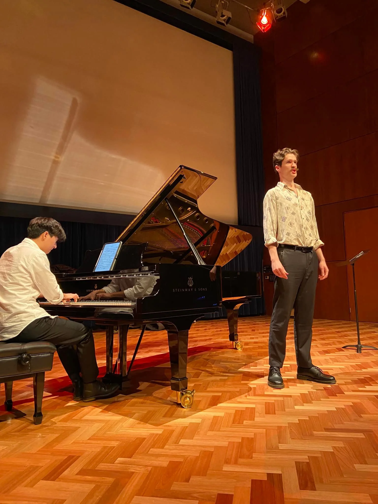 A stage with a person singing or speaking and a pianist playing a grand piano. The stage has a wooden floor and a large screen or backdrop behind them.