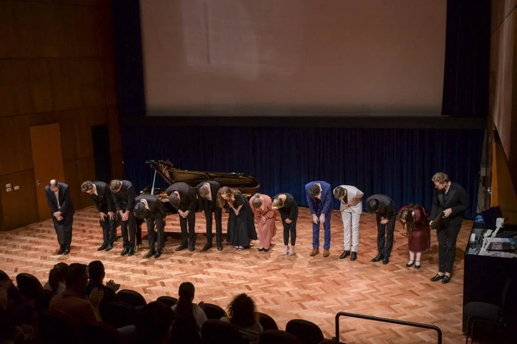 A group of people, including children and adults, standing on a stage bowing to an audience in a theater or concert hall.