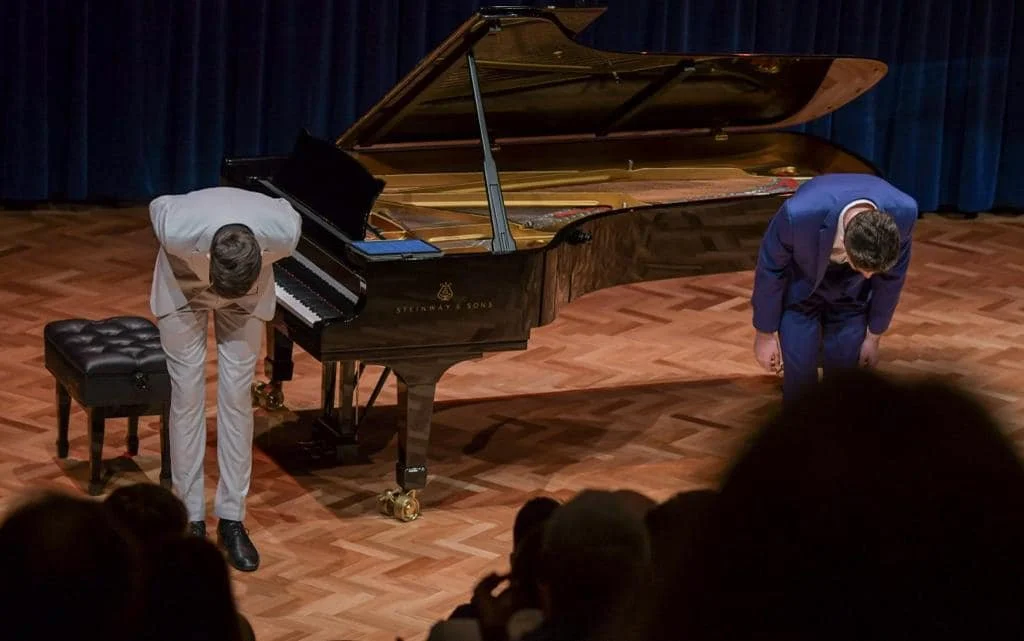 Two musicians bowing on stage after a performance, with a grand piano between them.