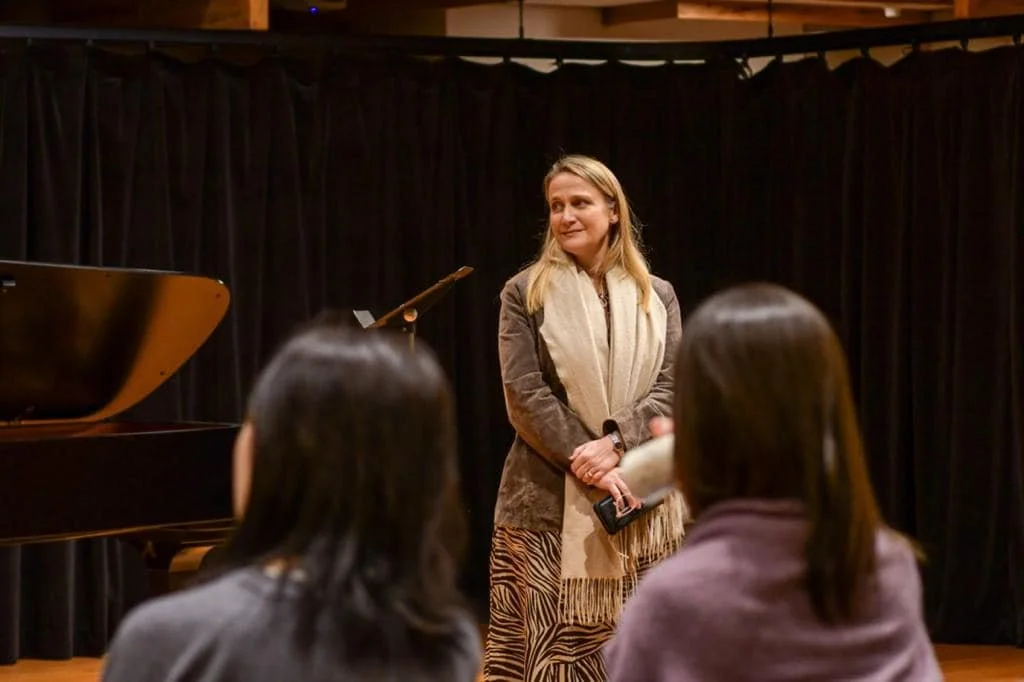A woman standing and speaking to an audience in a room with a black curtain background, a grand piano to her left, and people sitting and listening.