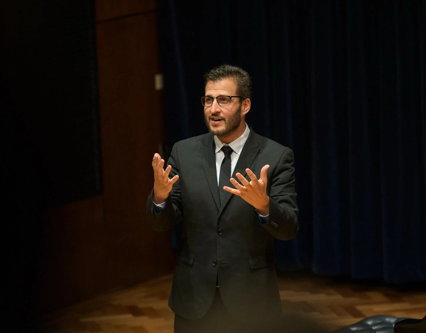 Man in black suit and glasses giving a speech or presentation in a room with dark curtains.