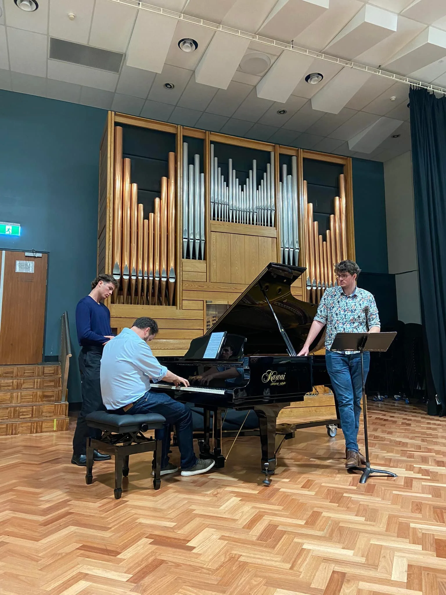 Three young men practicing a classical piano performance in a music hall. One is seated at a grand piano, while the other two stand nearby, one with a music stand, all engaged in the performance. A large organ is visible in the background.