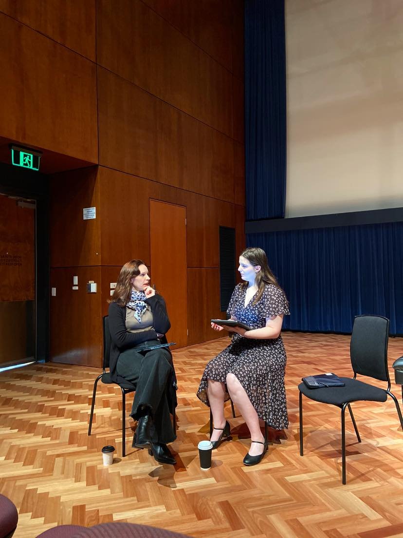 Two women sitting and talking in an auditorium or theatre with a wooden floor and blue curtains. One woman has dark hair and is wearing a black and beige outfit with a scarf; the other woman has light brown hair and is wearing a black floral dress, h
