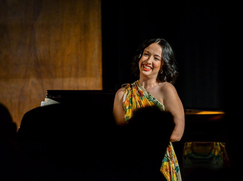 A woman with dark, wavy hair smiling on stage with a piano behind her.