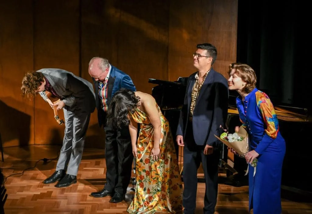 Five people standing on a stage bowing, with a wooden wall behind them and a grand piano to the right. The group includes three women and two men dressed in formal and colorful attire, with one woman holding a bouquet of flowers.