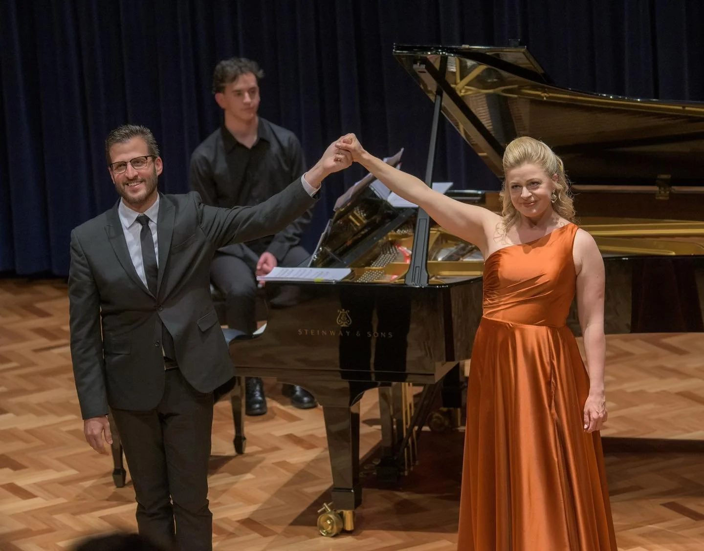 A man in a black suit and a woman in a satin orange gown are standing hand in hand in front of a grand piano on stage, with a pianist in the background.