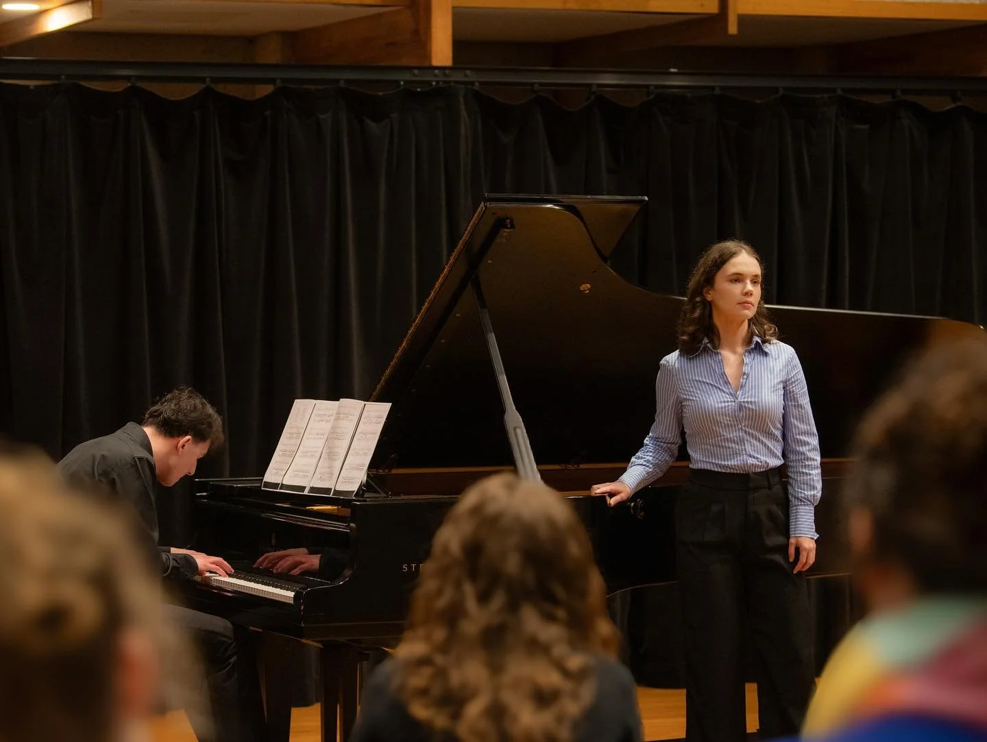 A woman stands at a grand piano on a stage while a man plays the piano. They are performing in front of an audience with black curtains behind them.