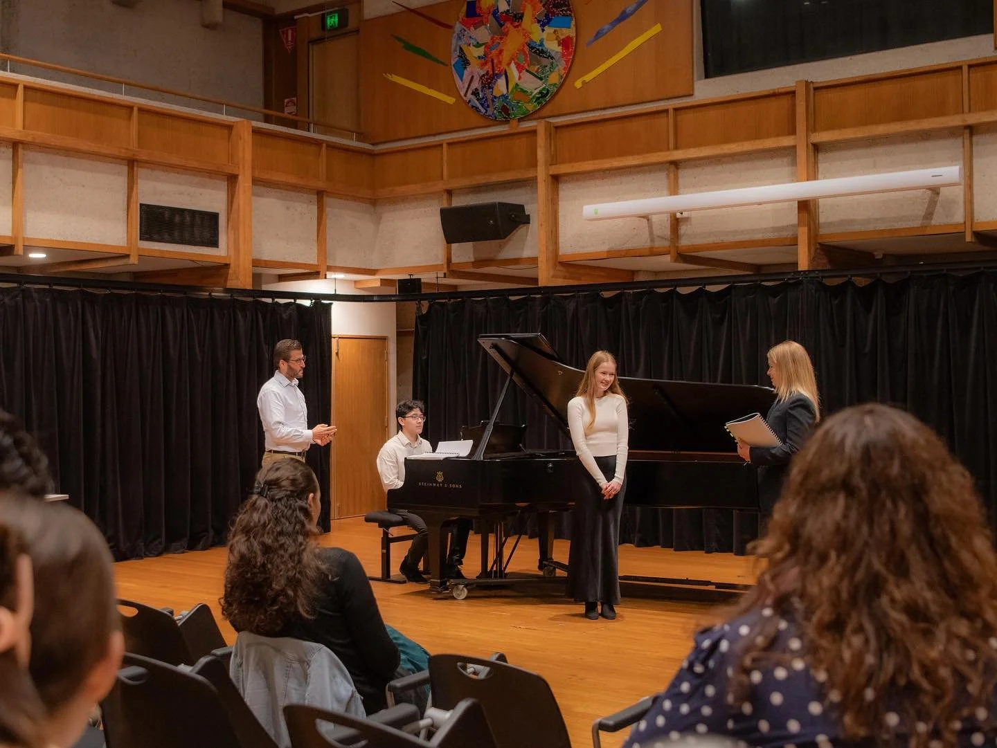 A musical performance on stage with a grand piano. A woman in a blazer is reading from a book, a girl is smiling at her, a man is standing, and a boy is seated at the piano. Audience members are seated in front. The setting is a room with wooden wall