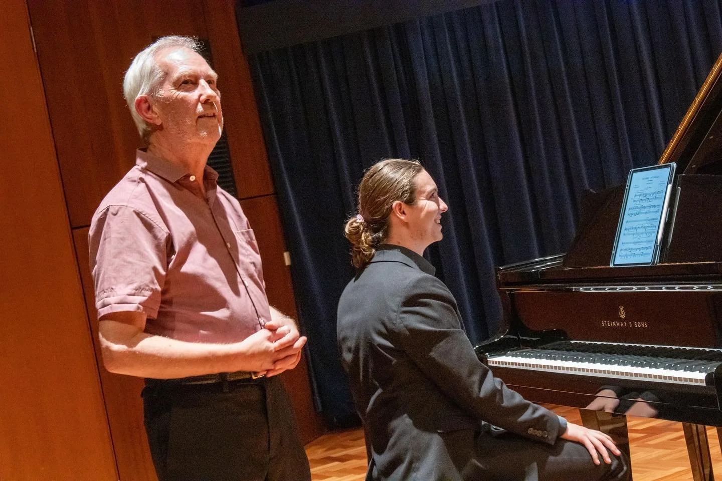 A man and a woman at a concert, the woman playing a grand piano and the man standing beside her.