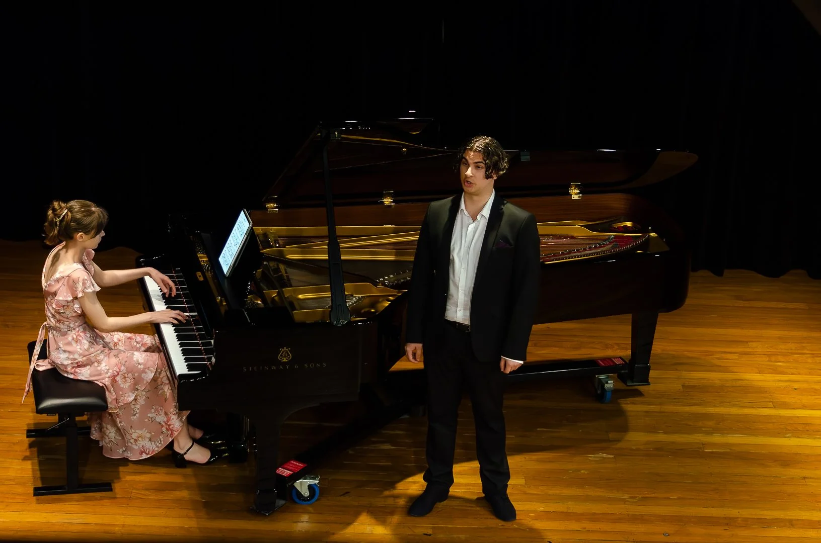 A young woman in a floral dress playing a grand piano on stage, with a man in a black suit singing beside her, in a concert hall setting.