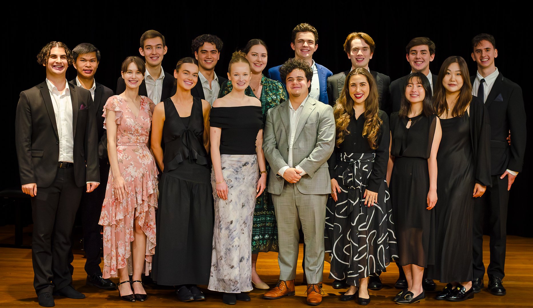 Group of young adults in formal attire standing on stage with black background, smiling.