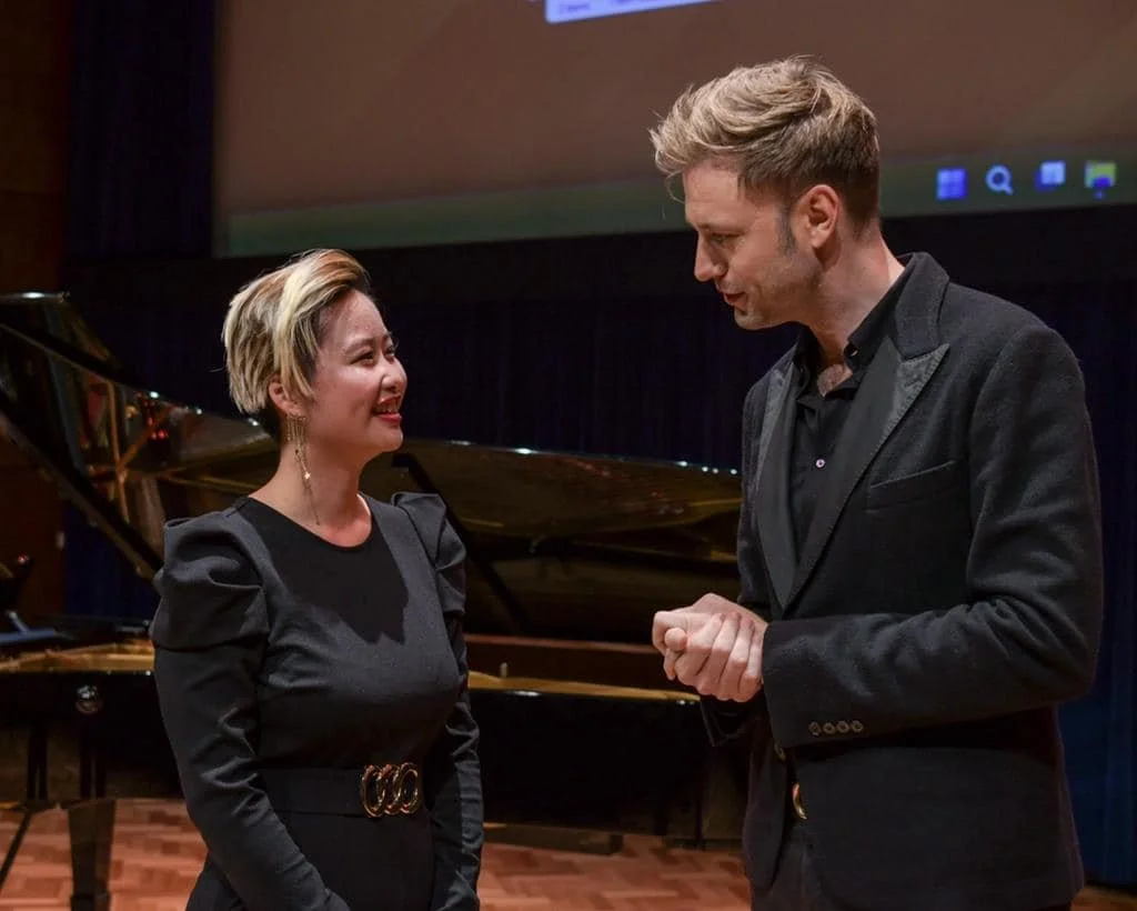 A woman and a man are standing and talking in a music venue with a grand piano behind them. The woman is smiling and wearing a black dress with puffed sleeves, while the man is looking at her and wearing a dark blazer.